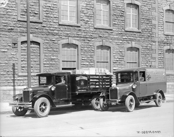 View across street towards two men in the driver's seats of two trucks with Quebec license plates. The sign painted on the side of the truck reads: "Teas and Coffees." There is a large stone building behind the trucks.