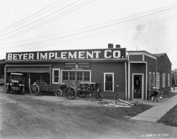 View across dirt road towards a dealership for International Harvester. There is a truck, Farmall tractor and a manure spreader on display in front of garage doors. Near the corner entrance near the sidewalk is a hand pump, and on display is a cream separator and milk cans.