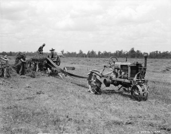 View across field towards a group of men using a Farmall to belt drive a hay press.