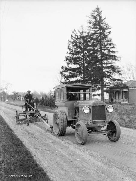A man is driving an industrial tractor up a road pulling another man behind him standing on a road grader. There is a house in the background on the right.