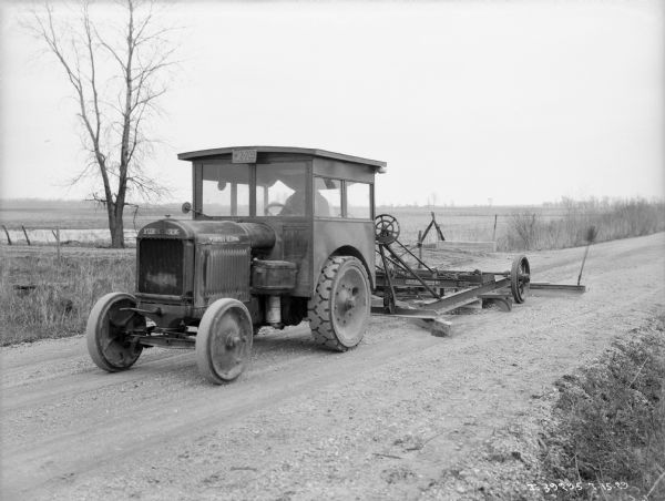 View towards a man driving an industrial tractor pulling a grader on a road. The sign painted on the grader reads, in part: "Adams Road __ntainer No. 6."