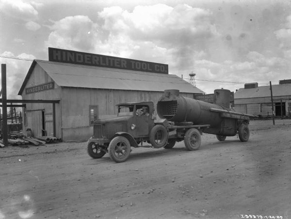 View across dirt road towards a man in the driver's seat of a truck. The truck is hauling a large section of pipe chained to rear wheels and resting on the open back section of the truck. In the background is an industrial building with a sign for "Hinderliter Tool Co." Other buildings and a tower are in the background.