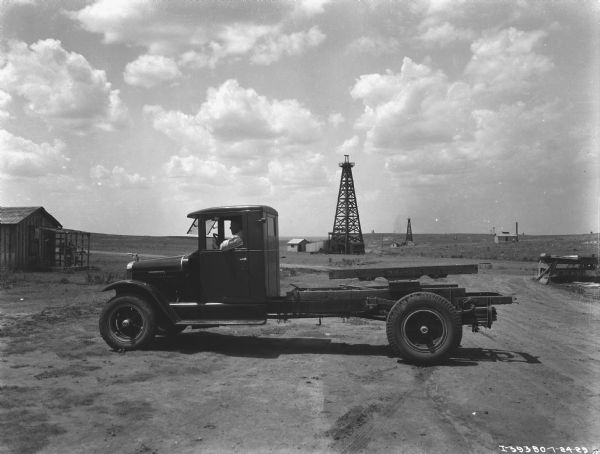 A man is sitting in the driver's seat of a delivery truck with an exposed truck bed. In the background are industrial buildings and oil wells.