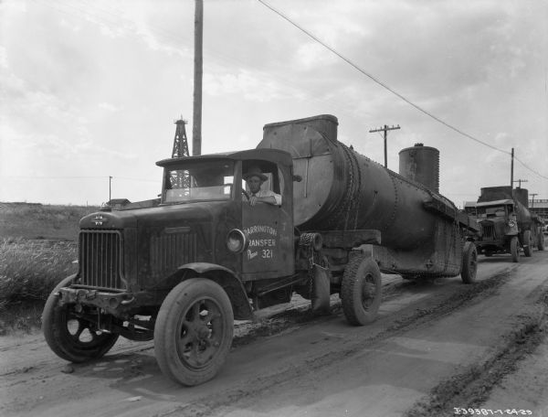 View across dirt road towards a man in the driver's seat of a delivery truck. The truck is hauling a large section of pipe chained to rear wheels and resting on the open back section of the truck. The sign painted on the driver's side door reads: "Barring Transfer." Behind the first truck is another man driving a truck with a similar section of pipe. There is an oil well in the background.