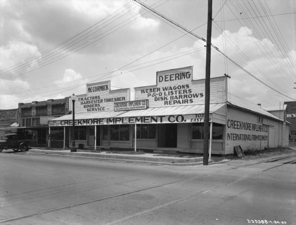 View from street towards a large dealership on a street corner. Railroad tracks are in the foreground. The sign on the awning of the building reads: "Creekmore Implement Co."