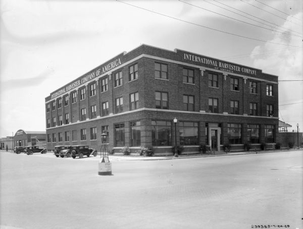 View across street towards a dealership. There are automobiles parked along the left side of the three-story brick building.