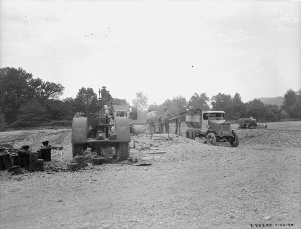 A group of men are working at a construction site near a dump truck. There is a tractor in the foreground providing belt-driven power. In the background on the left a man is driving a continuous track tractor.