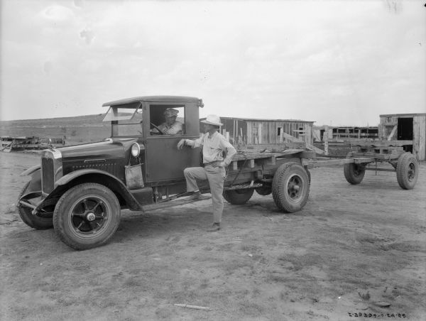 A man is standing near the driver's side door of a truck talking to a man sitting in the driver's seat of the truck. There are buildings in the background, and a bare hill in the far background.