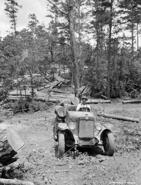 View towards front of International truck. A man is sitting in the driver's seat, and logs are in the truck bed behind him. The truck does not have a cab. Painted on the right side of the engine cover is: "No. 2." Felled logs are on the hill among trees in the background.