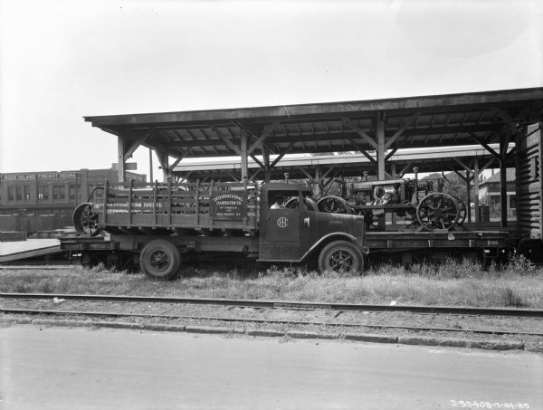 View across road and railroad tracks towards a man sitting in the driver's seat of a truck with a stake body. The sign on the side of the truck reads: "International Harvester Co. of America, Inc. 300 Ferry St." The truck is parked alongside a railroad car that has Farmall tractors on it. There is a platform with a roof behind the railroad car. In the background on the left is a brick building with a sign painted along the roof line that reads: "Rock Island Freight House."