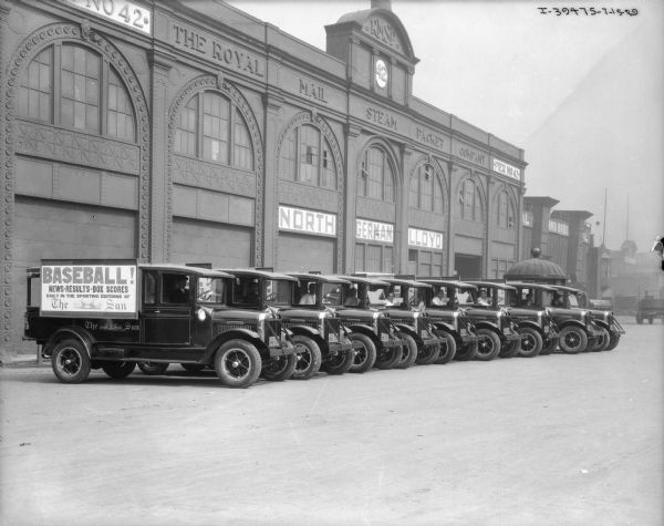 View across road towards men sitting in the driver's seats of a fleet of about nine delivery trucks parked in a row backed up against a large building. The sign along the top of the building reads: "The Royal Mail Steam Packed Company Pier No. 42." The sign on the side of the truck at the far left reads: "Baseball! News-Results-Box Scores Daily in the Sporting Editions of The Sun." The trucks have commercial license plates for New York.
