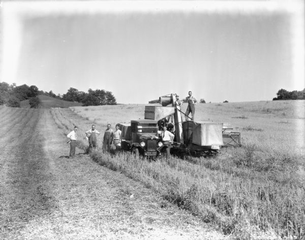 View across field towards a group of men posing with a truck and a McCormick-Deering harvester-thresher.