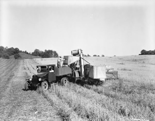 Slightly elevated view of a man sitting in the driver's seat of a truck, and another man sitting in the passenger seat. There is another man standing in the bed of the truck positioning a McCormick-Deering harvester-thresher. The field is partially harvested.