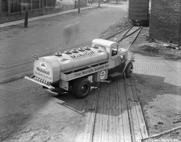 Elevated view of a Gargoyle Mobiloil delivery truck parked near two railroad tracks. The sign painted on the passenger side door reads, in part: "Vacuum Oil Company, __Dearborn St., __ago." A railroad car is sitting on the combined railroad tracks in the background near a brick building. Pedestrians (blurred from motion) are walking on the cobblestone street, and on the sidewalk in the background.