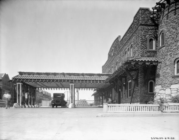 View towards a service station. The station is a large, brick building, with a roof over the pumps. A man is standing at the rear of an automobile filling the tank. Another man is sitting on a bench near the entrance on the right. A sign above the entrance reads: "Standard Oil Company Products."