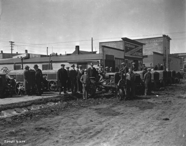 View down dirt road towards a group of men standing among Farmall and McCormick-Deering tractors in front of the J.W. Graham storefront. There is a boy sitting on a Farmall tractor in the center. There are other storefronts and a bank in the background on the right.