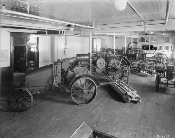 Slightly elevated view of tractors and agricultural implements on display at a dealership. An enclosed office area is in the back right corner.