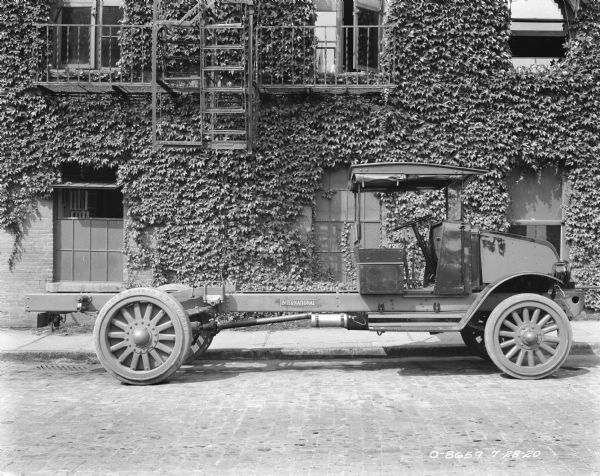Passenger side view of an International Harvester truck parked in front of a vine-covered brick building with a fire escape. The back of the truck has an exposed chassis. There is a running board, a bench seat for the driver and passenger, a windshield, and cab covers which are rolled up on three sides of the roof. The engine area is enclosed, and wide fenders are over the front wheels which have Goodyear tires.