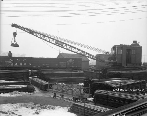 Elevated view of lumber stacked in a railroad yard. A man is standing inside a New York Central Lines railroad car in the background on the left. Two men are standing in the open doorway of the cab on a Brownhoist crane on the right. Snow is on the ground.
