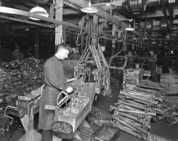 A man wearing a work apron is standing and working on an assembly line in a factory building. Other men are working in the background. In the far background in the center two men wearing hats and suits are watching. There are large piles of parts on the floor on the left and the right.