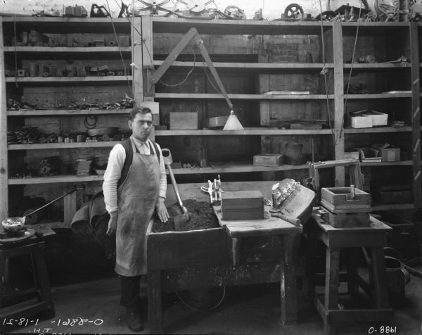 A man wearing a work apron is posing in a finishing room near a workbench. There is a shovel resting in dirt in a bin next to him, and cast parts are near wooden forms stacked on the right. On the far left is a crucible resting on a small bench. On the wall in the background are shelves of parts and other utensils.