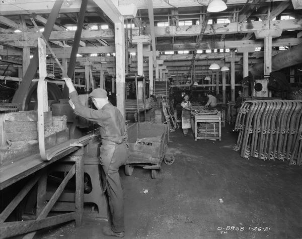 A man is working on assembling a part for a cultivator in a large factory area. Other men are working in the background. Parts are stacked on the right.