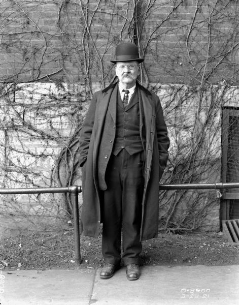 Full-length portrait of a man standing outdoors in front of a railing and brick factory wall. He is wearing eyeglasses, coat, suit and hat.