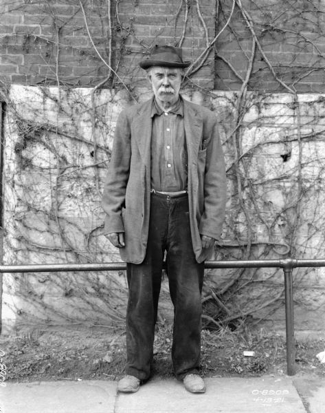 Full-length portrait of a man standing outdoors in front of a railing and brick factory wall.