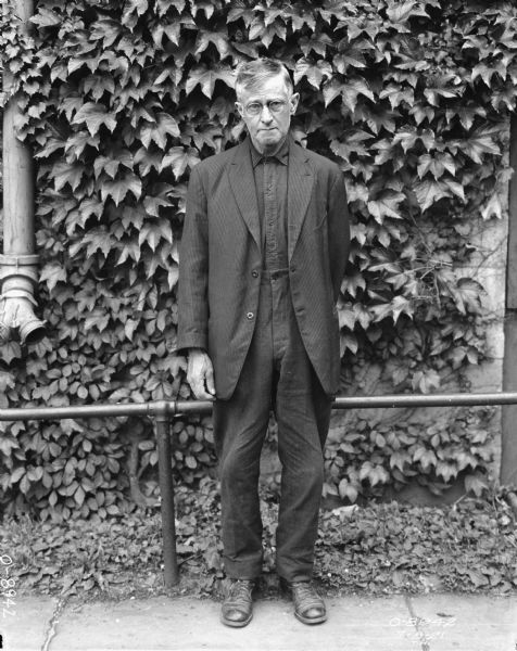 Full-length portrait of a man standing outdoors in front of a railing and brick factory wall covered with vines.