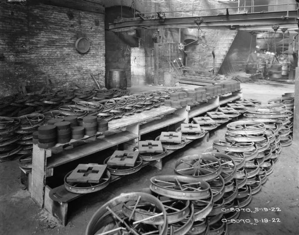 Wheels are stacked on the floor and on wood shelves. A flight of steps in the background is rising up to another story. Men (blurred from movement) are working in the background.