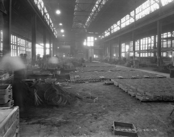 View down a large, open factory space towards a furnace oven. Parts are stacked on the floor. There are skylights in the ceiling.