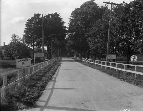 View down road with fences on both sides. Signs read: "Danger! NYH R.R. 300 Ft. Ahead," and "Incorporated Village of Minoa Slow Down to 15 Miles." There is an automobile parked near a house in the background on the left.