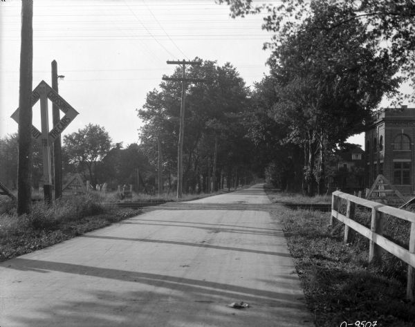 View down road with towards a railroad crossing. In the background on the right among trees is a brick building, and a house with a front porch.