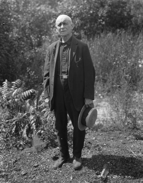 Full-length portrait of a man standing outdoors. He is wearing a suit jacked over a dark shirt and pants, and is holding a straw hat in hhis left hand. Plants and trees are in the background.