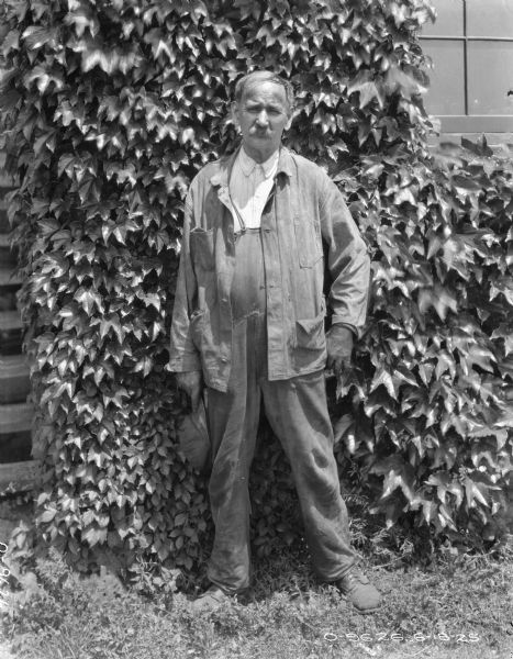 Full-length portrait of a man standing outdoors in front of a wall covered with foliage. He is holding a hat in his right hand.