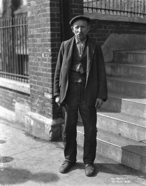 Full-length portrait of a man standing on a sidewalk. Behind him is a set of steps next to a brick wall with an iron fence. The man is wearing worn clothes, including a hat, suit jacket, vest, and torn pants.