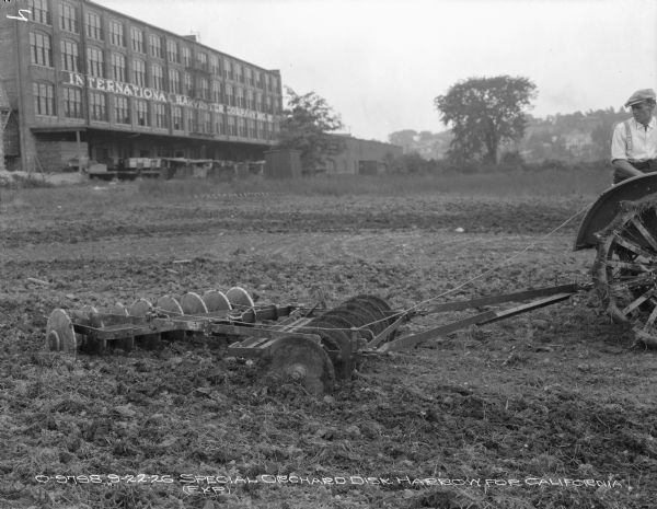 A man driving a tractor is using a disk harrow in a field. In the background is an IH plant.