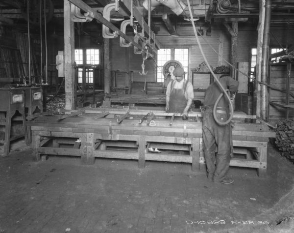 View of a factory area where two men are working. The man on the right has his back turned while using a hose attached to the ceiling. The other man is looking down at a part set up on a work table. Windows are along the back wall.