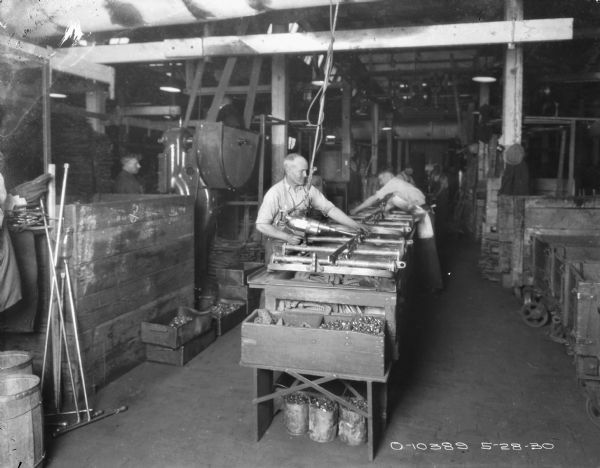 Men are working on parts set up on a worktable. The man in the foreground is using a tool with a long cord that is attached to the ceiling. In the background on the left a ma is standing near large machinery that is belt-driven.
