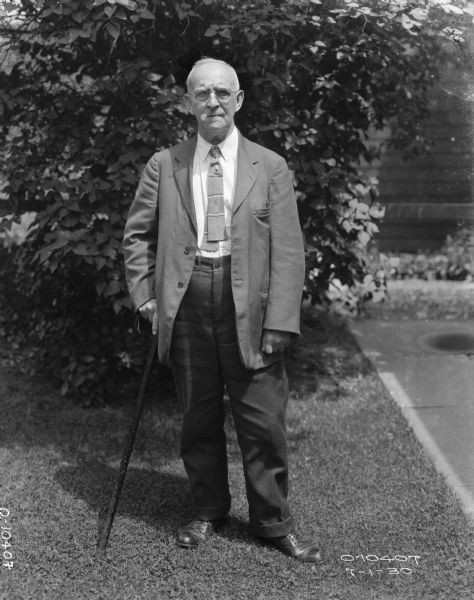 Full-length portrait of a man standing outdoors on a lawn near a sidewalk. He is wearing a suit, eyeglasses, and is leaning on a cane he is holding in his right hand. In the background are shrubs, and the brick wall of a building.