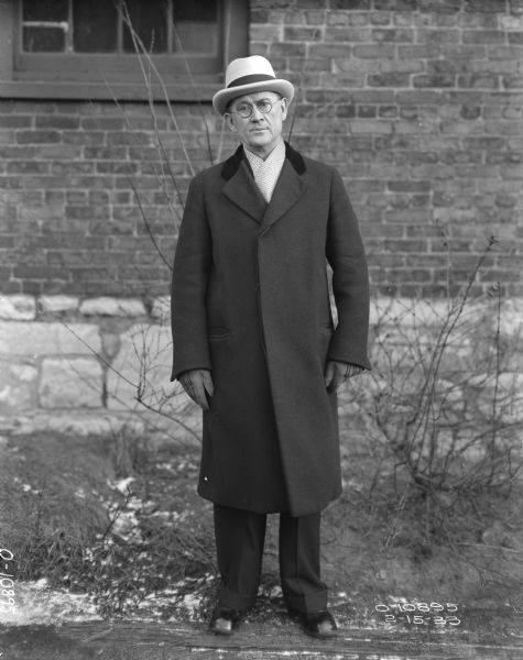 Full-length portrait of a well-dressed man standing outdoors. He is wearing a hat, eyeglasses, three-quarter length coat, gloves and a scarf. Behind him is the brick and stone wall of a factory building.