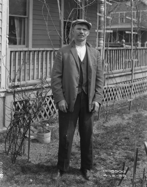 Full-length portrait of a male employee of Auburn Works standing in a yard. Behind him is a house with a porch.
