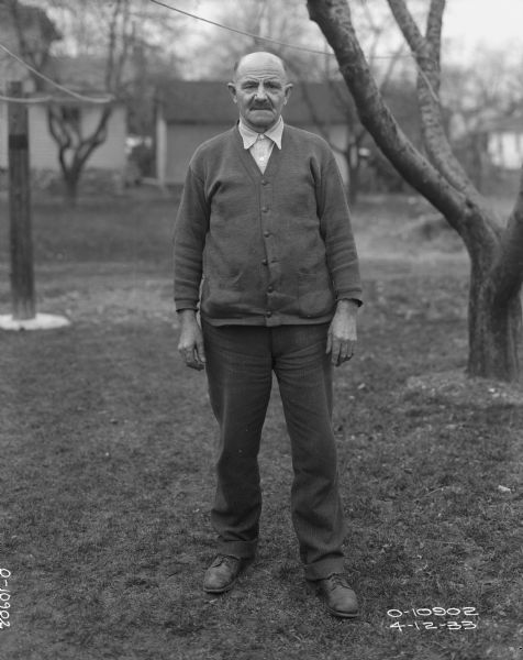 Full-length portrait of a male employee of Auburn Works standing outdoors on a lawn. He is wearing a cardigan over a button-down shirt. There are trees and buildings in the background.