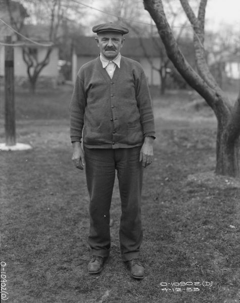 Full-length portrait of a male employee of Auburn Works standing outdoors on a lawn. He is wearing a hat, and a cardigan over a button-down shirt. There are trees and buildings in the background.