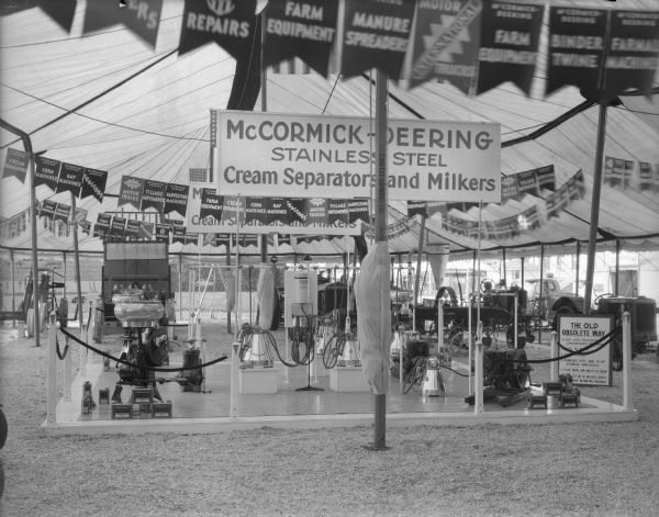 McCormick-Deering stainless steel cream separators and milers on display  on a platform underneath an open-sided tent. Tractors and other equipment are on display in the background.