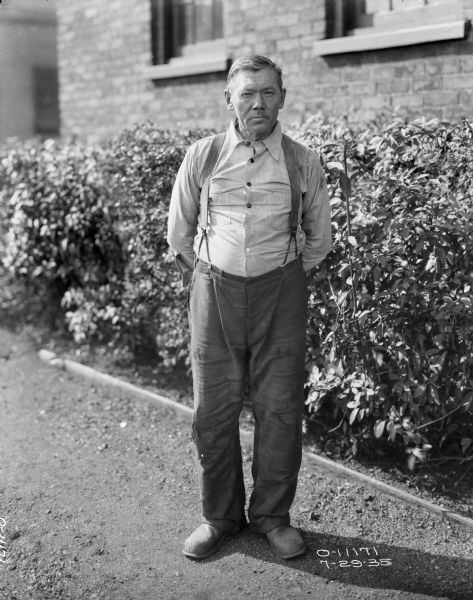 Full-length portrait of a man standing outdoors in front of bushes along the side of the brick wall of a factory building with windows. He is wearing work clothes and suspenders.