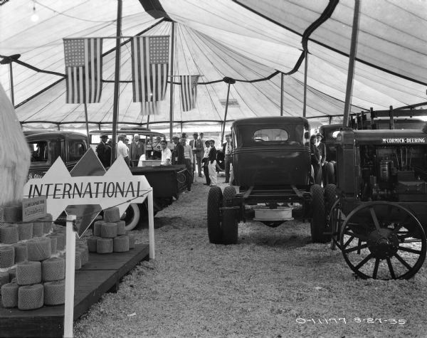 McCormick-Deering twine, trucks, and other equipment on display under an open-sided tent. Banners and flags are hanging above the equipment. Groups of men and boys are looking at the displays in the background.