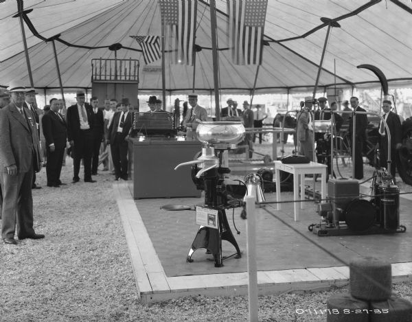 A cream separator is on display on a platform under a large, open-sided tent. Men are standing around looking at the displays. Flags are hanging above the display, and tractors and other equipment are in the background.