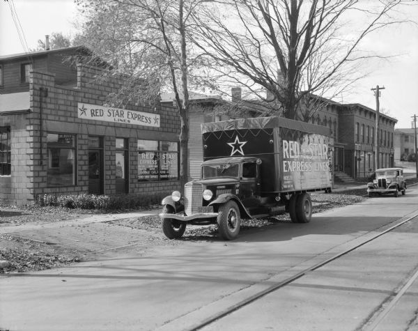 View across street towards a truck. The sign painted on the side of the truck reads: "Red Star Express Lines." The truck is parked along the curb in front of a commercial storefront for the Red Star company. Signs on a building further down the street are for International Harvester, and McCormick-Deering service.