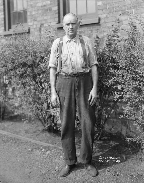 Full-length portrait of a man standing outdoors in front of bushes along the side of the brick wall of a factory building with windows. He is wearing work clothes and suspenders.